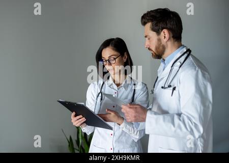 Doctors reviewing patient information on digital tablet and clipboard. Medical professionals collaborating on diagnosis. Healthcare teamwork for optimal patient care. Doctors wearing white lab coats Stock Photo