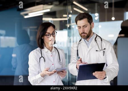 Two doctors in white coats reviewing patient data on a clipboard and tablet in contemporary medical office. Focused healthcare professionals collaborating during work meeting Stock Photo