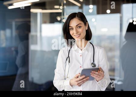 Professional female doctor smiling while using digital tablet in modern medical office. Confident healthcare worker managing patient records and healthcare services. Stock Photo