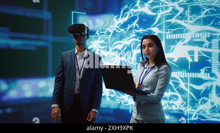 Technician wearing VR headset looks at hologram of artificial intelligence human brain simulation, assisted by colleague using laptop. Team of programmers interacting with AI visualization, camera B Stock Photo