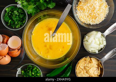 Beaten Eggs in a Glass Mixing Bowl with a Fork: Eggs for migas surrounded with tortilla chips, cheese, cilantro, onions, and jalapenos Stock Photo