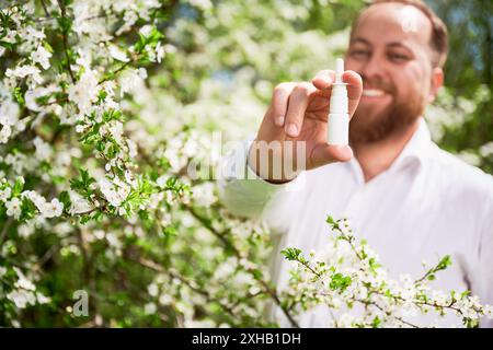 Focus on man hand's holding nasal spray. Smiling man allergic using medical nasal drops, suffering from seasonal allergy in blossoming garden. Spring allergy concept. Stock Photo