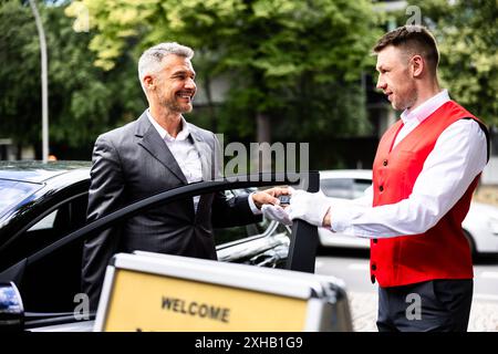 Luxury Car Rental Service: Professional Businessman Smiling as Valet Hands Over Keys beside Stylish Vehicle on Sunny City Street. Stock Photo