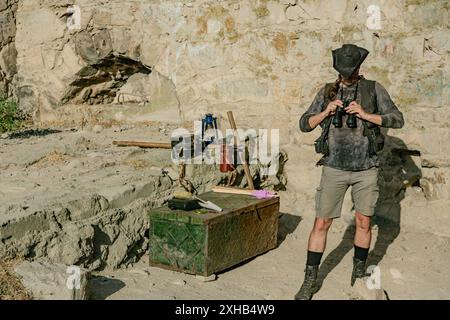 An archaeologist of the last century looks through binoculars against the background of a fortress wall and his equipment with a chest. Educational pr Stock Photo