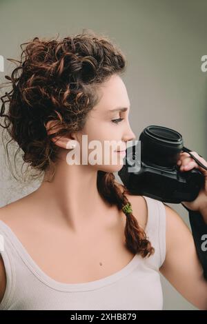 Woman with curly hair in a braid, wearing a sleeveless top, holds a camera close to her face. Her serene expression and slight smile reflect a calm an Stock Photo