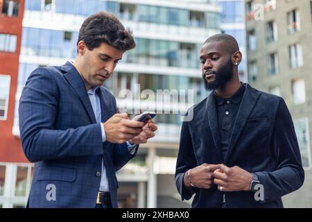Two businessmen, one black African American and one Caucasian, in business suits focused on their smartphone while standing in the financial district Stock Photo