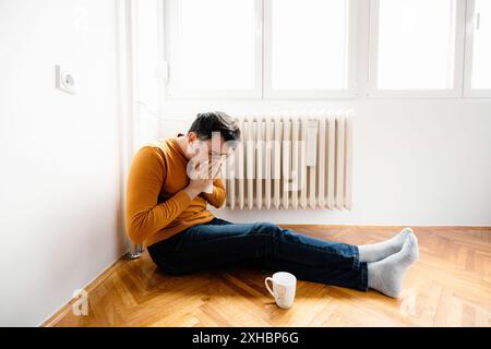 A man sits on the floor, wearing jeans and a yellow sweater, next to a radiator. He is holding a tissue and sneezing. There is a cup on the floor next Stock Photo