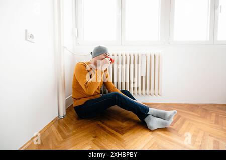 A man sits on a hardwood floor in a room with white walls, wearing a gray hat and a yellow sweater. He drinks from a red mug while looking to the side Stock Photo