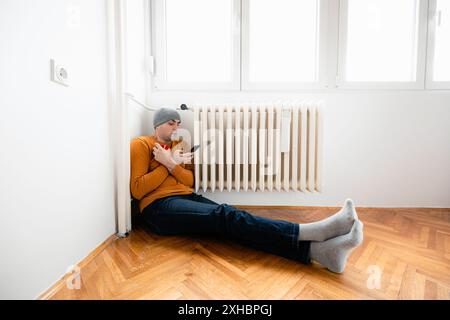 A man sits on the floor near a radiator, wearing a hat and warm clothing. He holds a mug in one hand and looks at his phone. Stock Photo