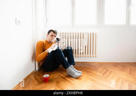 A man sits on the floor, leaning against a wall, next to a radiator. He is looking at his phone and holding a white napkin. Stock Photo