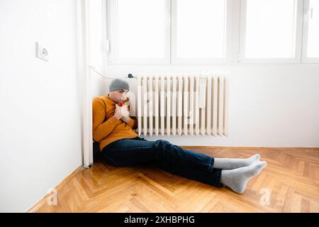 A man sits on the floor in a cold room, wearing a hat and an orange sweater, with his legs stretched out near a radiator. Stock Photo