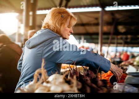 A woman with red hair carefully selects beets from a stall at an outdoor market. Stock Photo