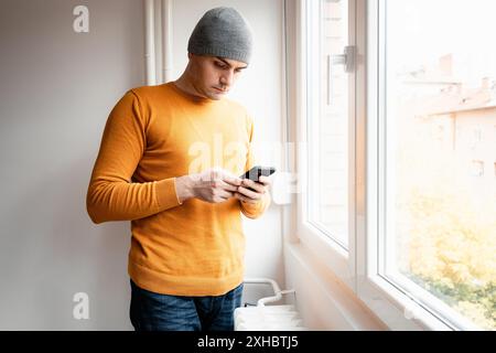 A man in a grey beanie and orange sweater uses his smartphone while standing next to a window. Stock Photo