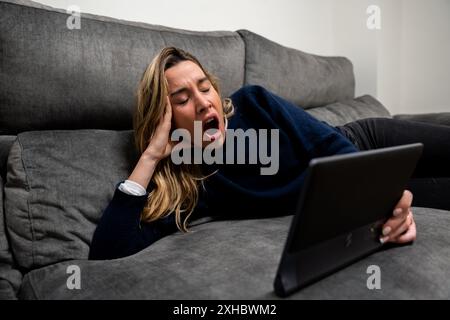 Woman lying on the sofa yawning with a tablet in her hand. Facial expression of sleep or boredom Stock Photo