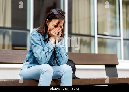 A woman sits on a bench outside a building, crying and holding her face. She is wearing a blue shirt and jeans. Stock Photo