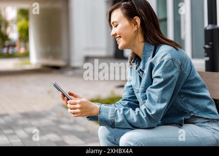A woman sits on a bench outdoors, smiling as she uses her smartphone. Stock Photo