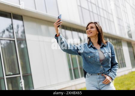 A woman takes a selfie with her smartphone while walking in front of a modern building on a sunny day. Stock Photo