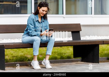 A young woman sits on a bench outside, smiling as she looks down at her phone. Stock Photo