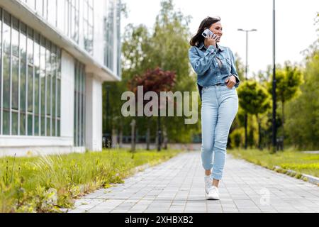 A young woman wearing a denim shirt and jeans walks down a sidewalk, smiling and talking on her phone. Stock Photo
