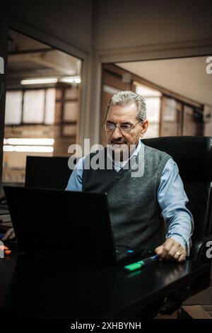 A mature man in a gray vest and blue shirt works on his laptop while sitting at his desk in an office. Stock Photo