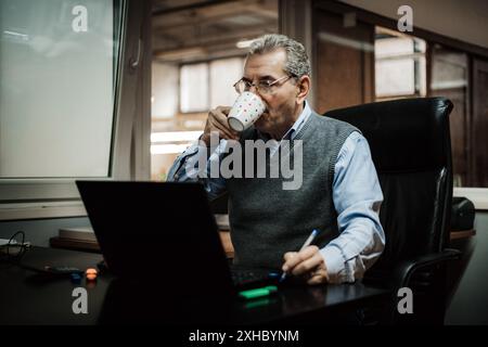 A man sits at a desk in an office, drinking coffee from a mug and working on a laptop. Stock Photo