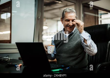 A senior man with gray hair laughs while working on his laptop in an office, holding a cup of coffee. Stock Photo