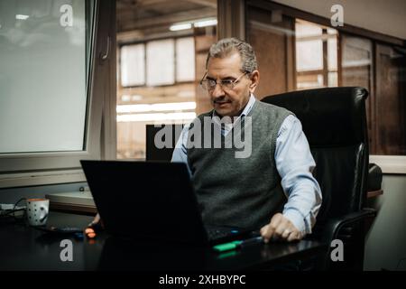 A mature man sits in an office chair at a desk, using a laptop. Stock Photo