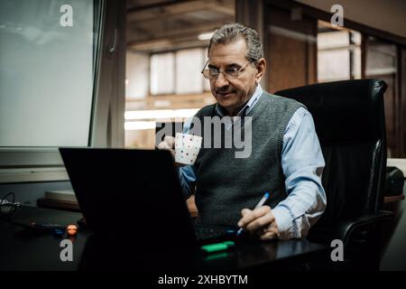 A man sits at a desk in an office, working on a laptop and holding a coffee mug. He is wearing a blue shirt and a grey sweater vest. Stock Photo