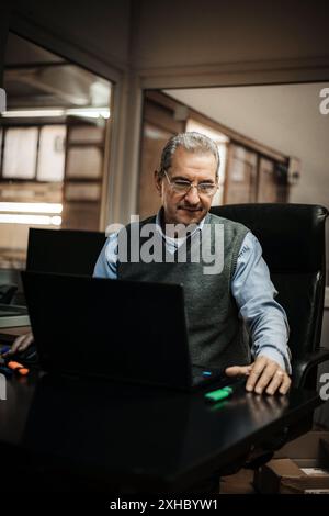 A man sits at a desk in an office, working on his laptop. Stock Photo