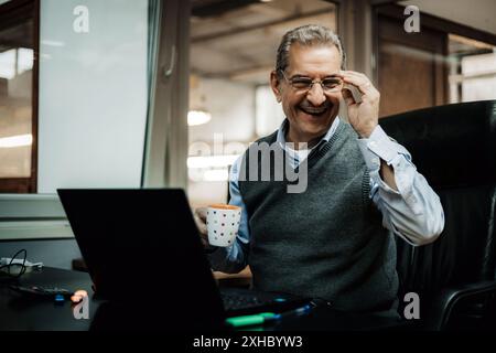 A man is seated at a desk in an office, laughing at a laptop while holding a colorful mug. Stock Photo