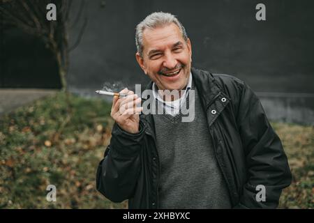 A man with grey hair wearing a black jacket and a grey sweater is smoking a cigarette. He is smiling and looking at the camera. Stock Photo