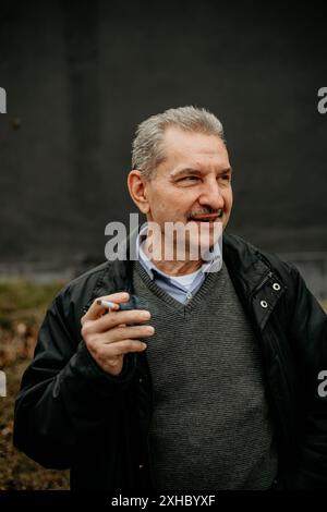 A middle-aged man with gray hair stands in front of a gray wall while holding a cigarette. Stock Photo