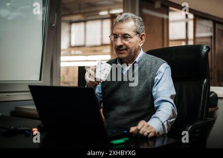 A senior man in an office setting, sitting at a desk, using a laptop while drinking coffee. Stock Photo