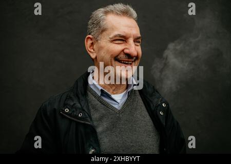 A man with gray hair and a mustache is laughing, with smoke exhaling from his mouth. He wears a black jacket over a gray sweater and a light blue shir Stock Photo