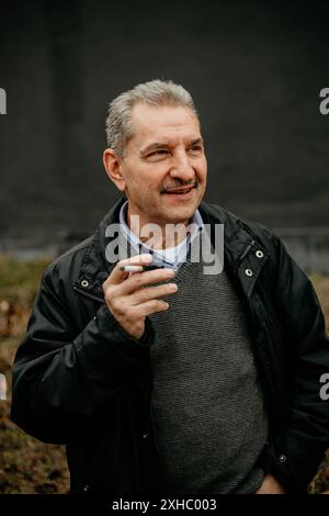 An older man, wearing a black jacket and a grey sweater, stands outdoors with a cigarette in his hand. Stock Photo