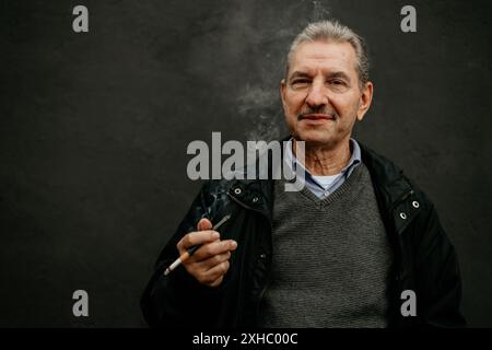 A man wearing a black jacket and gray sweater holds a cigarette and exhales smoke against a dark background. Stock Photo