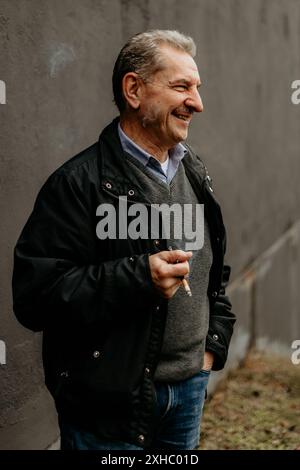 A man in a black jacket, gray sweater, and blue jeans stands in front of a grey wall, smiling and holding a cigarette. Stock Photo
