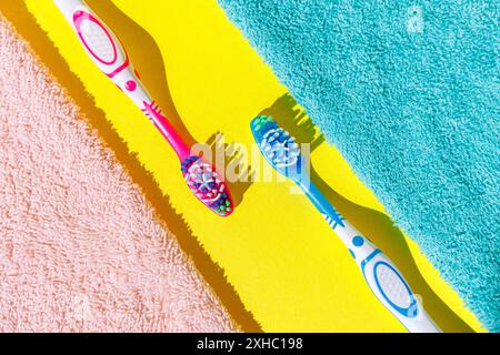 Two toothbrushes, one pink and one blue, lay on a bright yellow surface, each bordering a different colored towel. Stock Photo