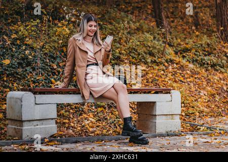 A woman sits on a bench in a park, surrounded by fallen leaves, looking at her smartphone. Stock Photo