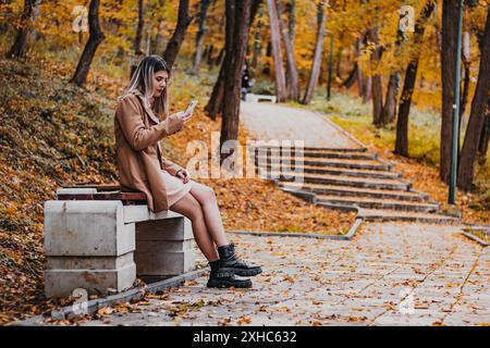 A young woman sits on a concrete bench in a park during autumn, looking at her smartphone. Stock Photo