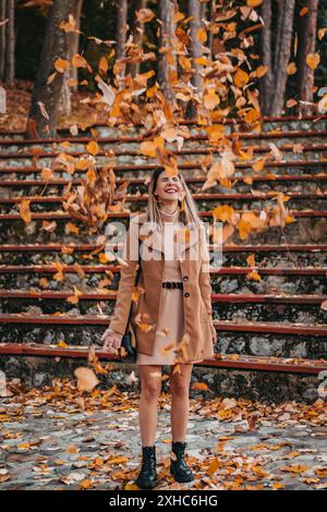A woman in a brown coat and dress stands on stone steps smiling as leaves fall around her. Stock Photo