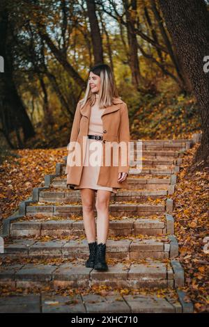 A young woman wearing a brown coat and a light dress walks down a stone staircase in a wooded area. Fallen leaves cover the ground and steps. Stock Photo