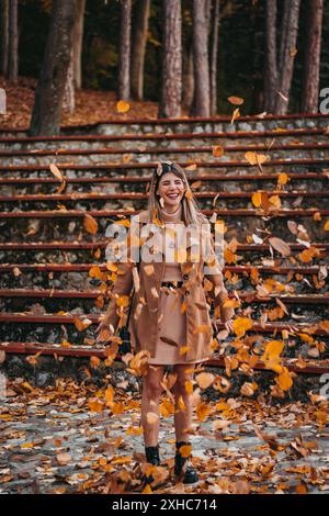 A woman in a brown coat stands in a forest, smiling as leaves fall around her. Stock Photo