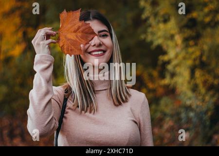 A young woman with blonde hair smiles while holding a large orange leaf in front of her face. The background is blurred autumn foliage. Stock Photo