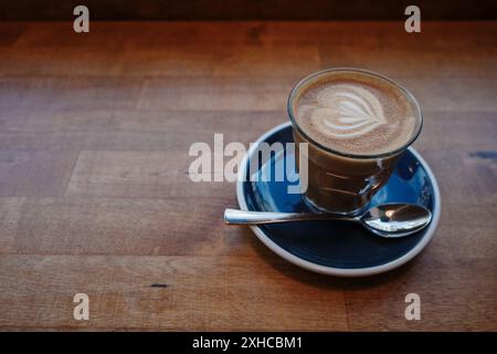 A beautifully crafted latte with intricate foam art in a glass cup, set on a dark blue saucer with a silver spoon. Stock Photo
