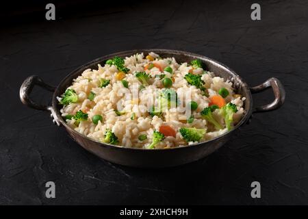 Vegan rice with vegetables in a pan, on a black background. Broccoli, green peas, and carrots in a healthy dish Stock Photo