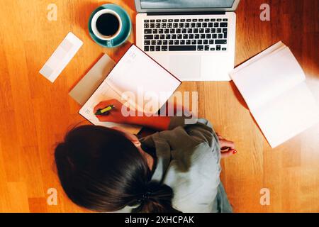 Top view of anonymous woman lying on floor near laptop and cup of coffee and writing in planner while working on remote project Stock Photo