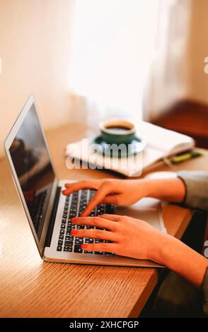 Anonymous female freelancer typing on laptop while working remotely from home at wooden table with books Stock Photo