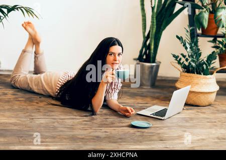 Calm female entrepreneur lying on wooden floor at home with laptop and drinking hot beverage while working remotely and looking at camera Stock Photo