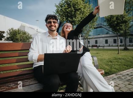Smiling colleagues working outdoors on a bench with a laptop and notebook on a sunny day Stock Photo